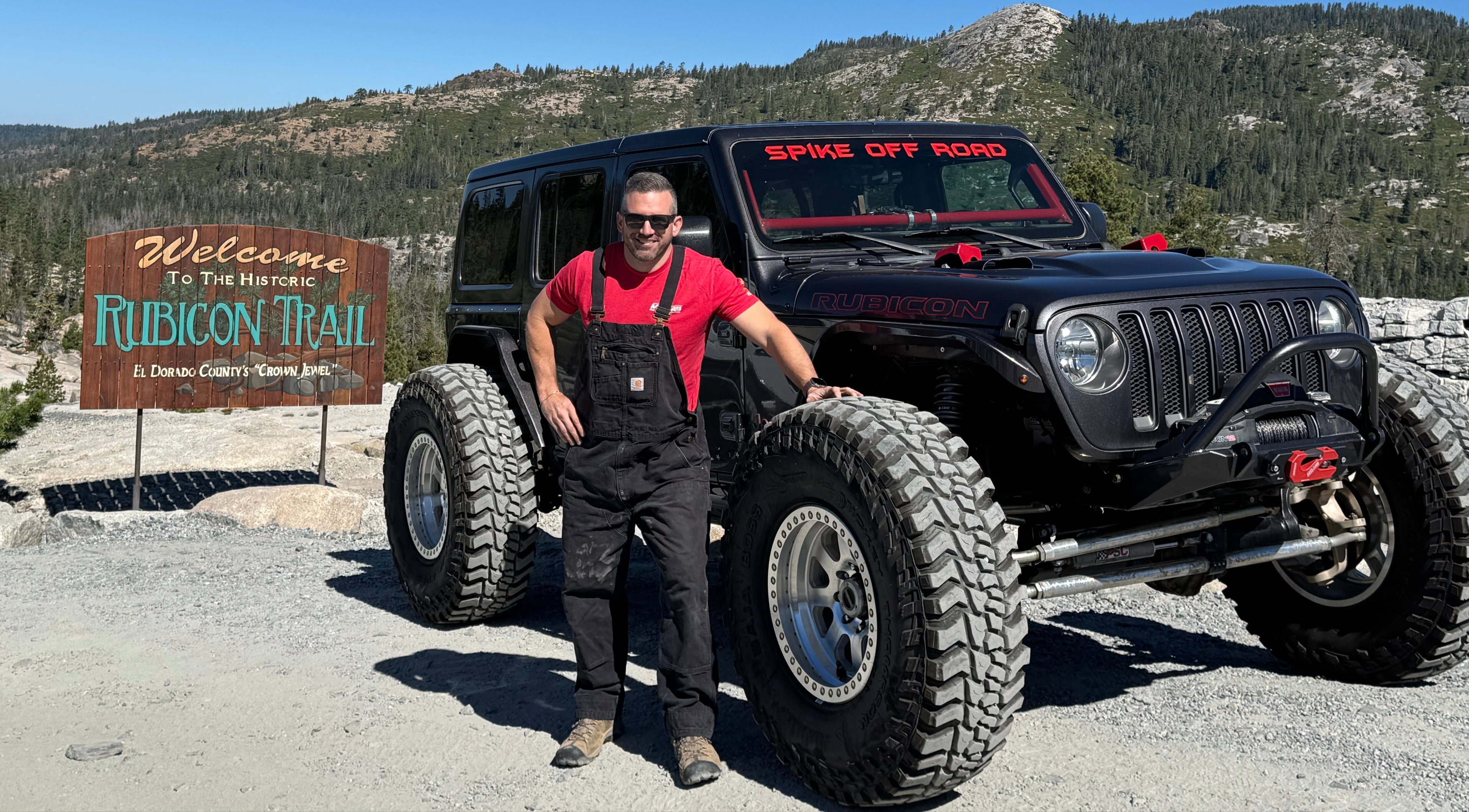 Man standing next to a black Jeep Wrangler at the Rubicon Trail entrance sign.